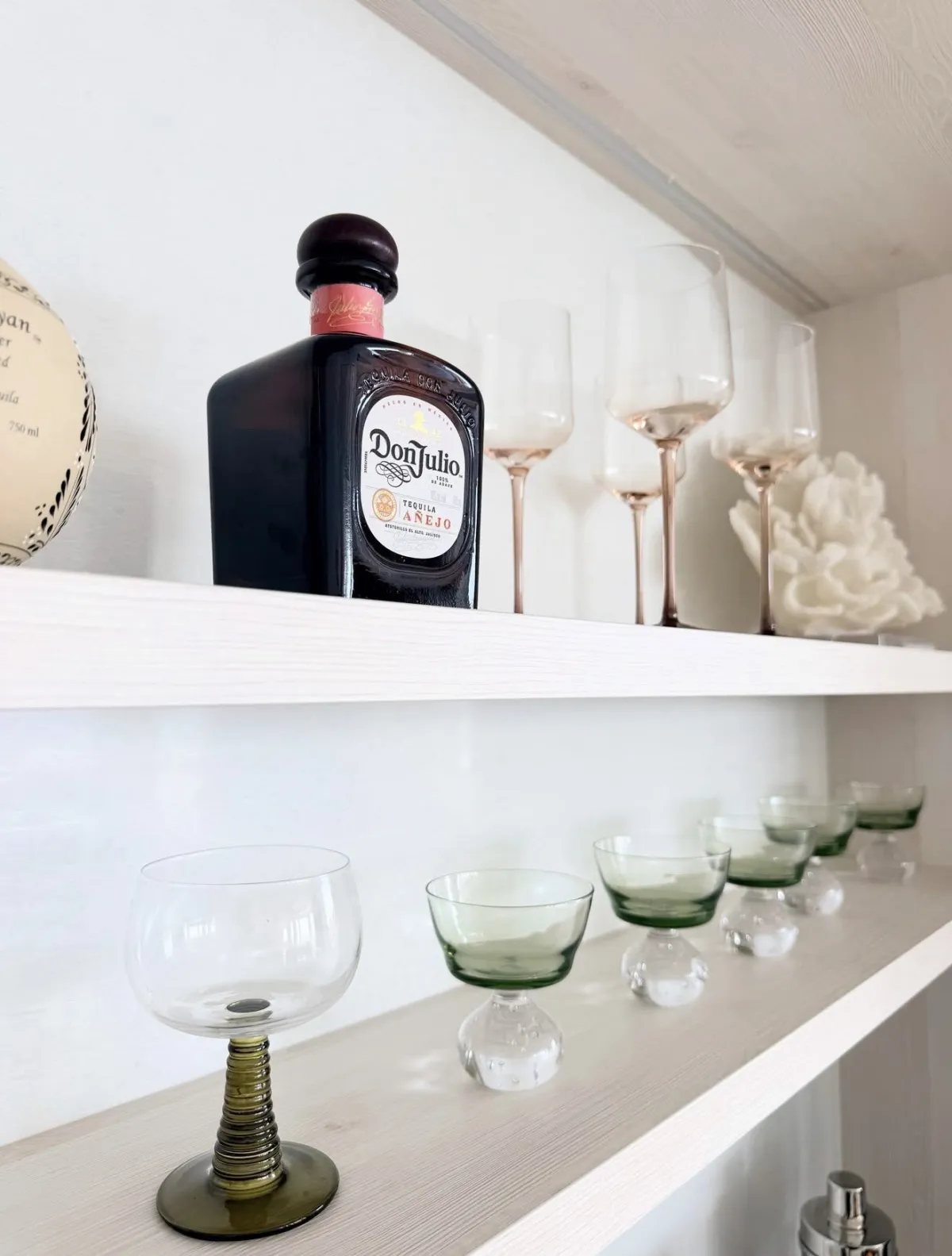 Don Julio bottle with sculptural stemware and green cocktail glasses on white bar shelves