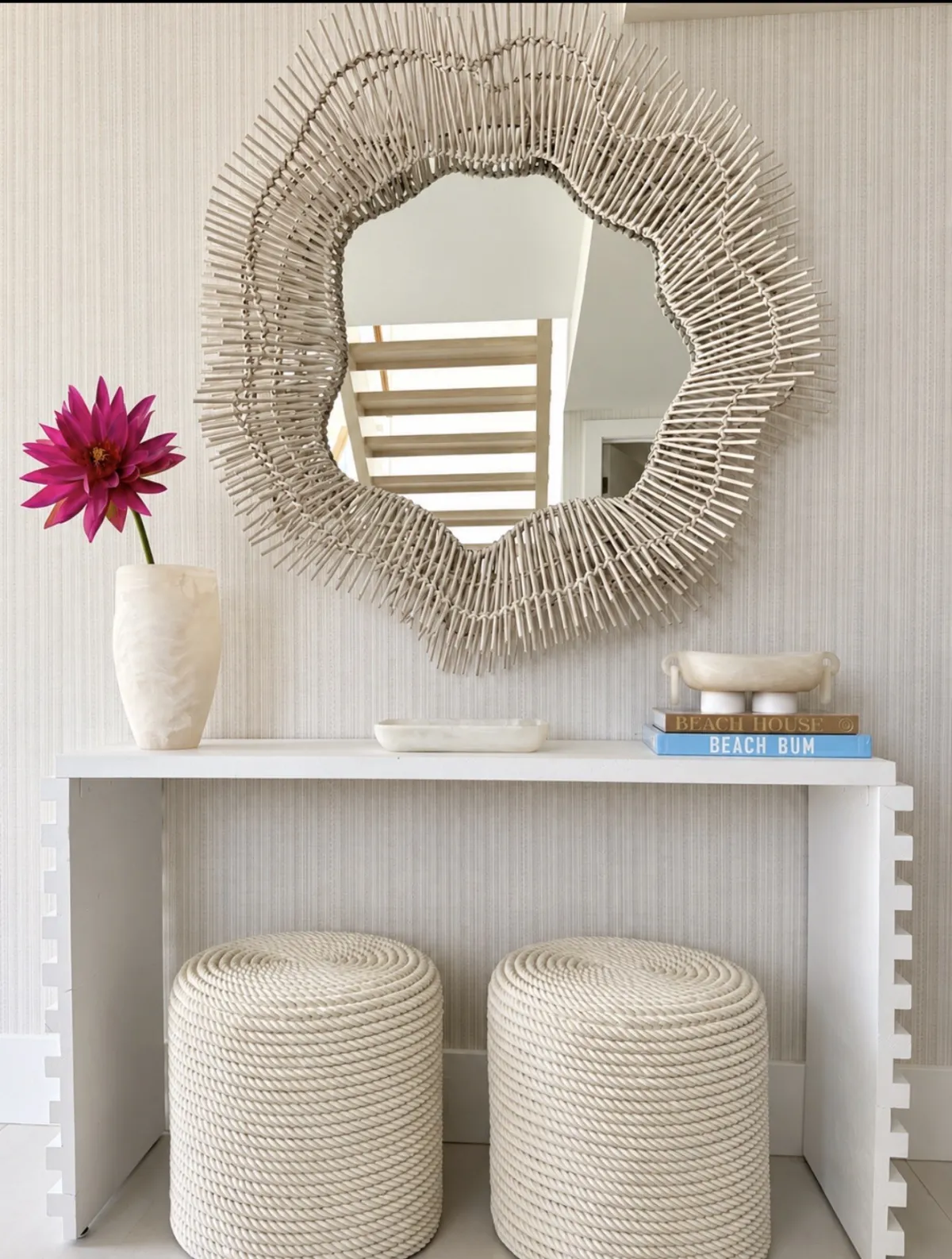 Woven sculptural mirror above a white entry console with magenta flower and two rope stools