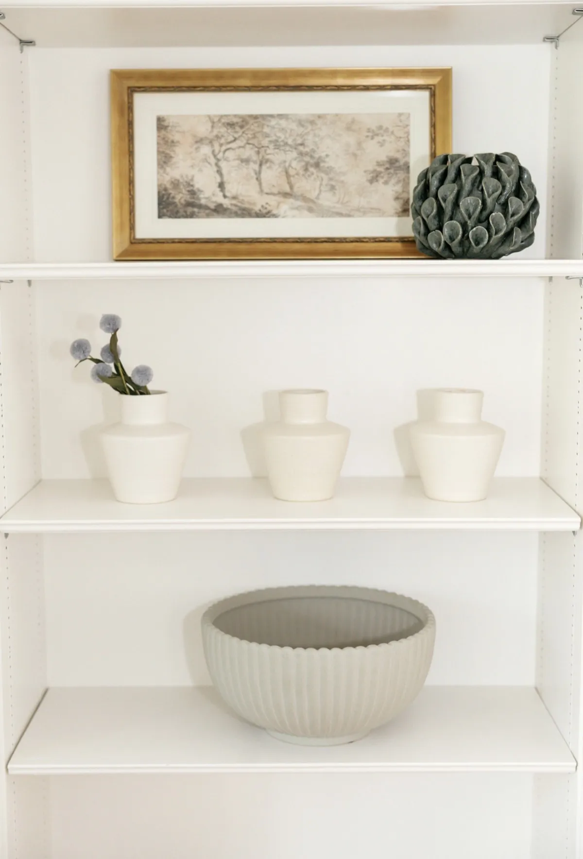 Framed landscape art, blue flowers, and a sculptural bowl on white shelves